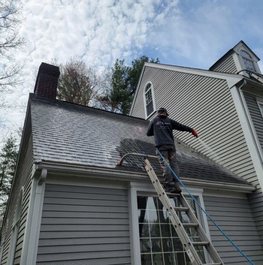 roof washing professional standing on ladder cleaning roof of gray home